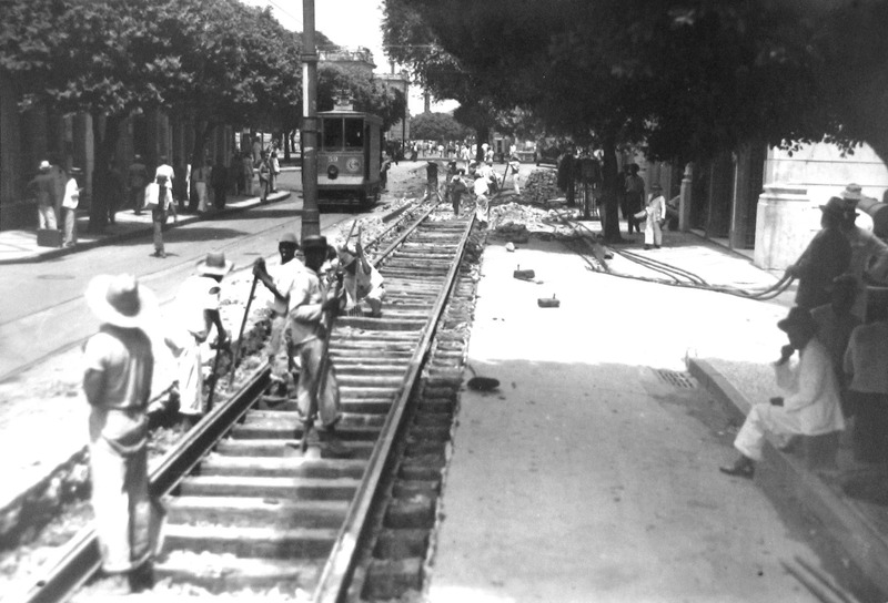 Rua com um bonde e trabalhadores negros construindo uma linha ferroviária